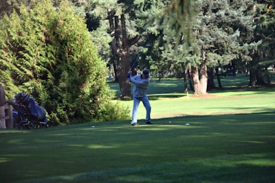 A person is preparing to swing a golf club on a lush, green golf course surrounded by tall trees. There is a golf bag with clubs nearby, resting on a cart. The lighting suggests it is a sunny day.