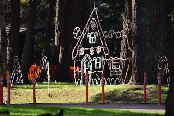 A large gingerbread house structure made of illuminated wire is displayed in a wooded park area. Candy cane decorations surround it, and orange safety barriers and poles are seen in the foreground. Sunlight filters through the trees, casting shadows on the grass.