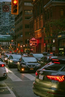 A busy urban street scene features numerous cars stuck in traffic under a red traffic light. The buildings are densely packed, with Massey Hall prominently displayed with its illuminated sign. The scene is set in the evening, casting a warm glow over the city environment.