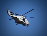 A military helicopter with two rotors is flying against a clear blue sky. The helicopter is painted in a light gray color and is equipped with a visible landing gear and other military equipment.