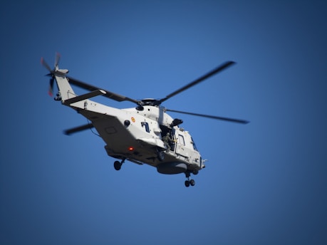 A military helicopter with two rotors is flying against a clear blue sky. The helicopter is painted in a light gray color and is equipped with a visible landing gear and other military equipment.