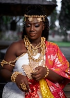 A person dressed in traditional attire with intricate gold jewelry, including necklaces, bracelets, and a headdress. The garment is adorned with vibrant colors like red, yellow, and white, featuring detailed patterns. The setting appears to be outdoors with blurred greenery in the background.