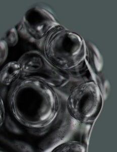 A close-up view of transparent bubbles clustered together with various sizes, creating a distorted and textured surface. The bubbles exhibit reflections and refractions of light, giving them a glossy and somewhat metallic appearance.