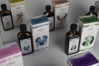 Several bottles and boxes of a product labeled 'UMbra' are arranged on a table. Each box and bottle features distinct artwork and numbers, with illustrations of various animals in different colors. The colors of the packaging are predominantly earthy and muted, with shades of blue, green, and brown. The boxes and bottles appear sleek and are likely part of a set or collection.