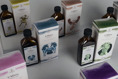 Several bottles and boxes of a product labeled 'UMbra' are arranged on a table. Each box and bottle features distinct artwork and numbers, with illustrations of various animals in different colors. The colors of the packaging are predominantly earthy and muted, with shades of blue, green, and brown. The boxes and bottles appear sleek and are likely part of a set or collection.