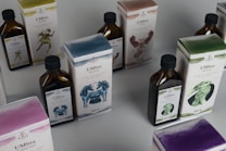 Several bottles and boxes of a product labeled 'UMbra' are arranged on a table. Each box and bottle features distinct artwork and numbers, with illustrations of various animals in different colors. The colors of the packaging are predominantly earthy and muted, with shades of blue, green, and brown. The boxes and bottles appear sleek and are likely part of a set or collection.