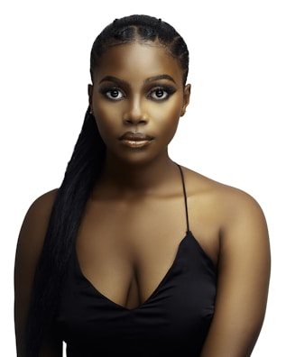 A woman with long braided hair styled in a ponytail wears a black spaghetti strap top against a plain white background. Her makeup is bold, featuring prominent eyeliner and strong contouring.
