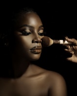 A close-up portrait of a person with flawless makeup, featuring glossy lips and well-defined eyebrows. A hand is applying makeup with a brush, highlighting the contours of the face. The lighting creates dramatic shadows, enhancing the details and texture of the skin.