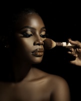 A close-up portrait of a person with flawless makeup, featuring glossy lips and well-defined eyebrows. A hand is applying makeup with a brush, highlighting the contours of the face. The lighting creates dramatic shadows, enhancing the details and texture of the skin.