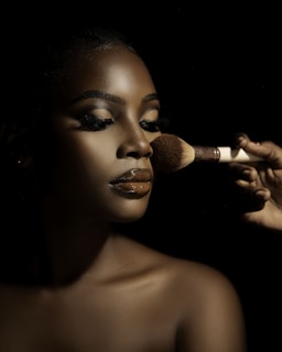 A close-up portrait of a person with flawless makeup, featuring glossy lips and well-defined eyebrows. A hand is applying makeup with a brush, highlighting the contours of the face. The lighting creates dramatic shadows, enhancing the details and texture of the skin.