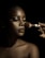 A close-up portrait of a person with flawless makeup, featuring glossy lips and well-defined eyebrows. A hand is applying makeup with a brush, highlighting the contours of the face. The lighting creates dramatic shadows, enhancing the details and texture of the skin.