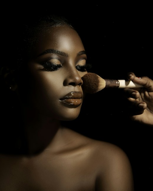 A close-up portrait of a person with flawless makeup, featuring glossy lips and well-defined eyebrows. A hand is applying makeup with a brush, highlighting the contours of the face. The lighting creates dramatic shadows, enhancing the details and texture of the skin.