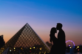 A romantic silhouette of a couple sharing an intimate moment in front of a large glass pyramid structure, likely the Louvre Pyramid. The background is a gradient sky transitioning from blue to orange, indicating either sunrise or sunset. A Ferris wheel is noticeable in the right portion of the image, with city lights dotting the scene.
