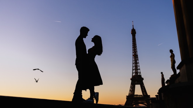 Silhouetted couple embracing against a backdrop of the Eiffel Tower during sunset, with birds flying nearby and a clear sky transitioning from orange to blue.