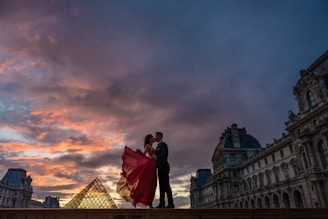 A couple stands romantically posed against a dramatic sunset sky, with the iconic glass pyramid and classical architecture creating a dynamic backdrop. The woman's red dress billows gracefully, complementing the warm hues of the sunset.