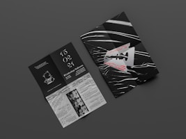 Two graphic print materials rest on a dark gray surface. One appears to be a folded booklet featuring bold red text and an abstract razor blade design with white and black wavy lines. The other is an unfolded brochure with information, elegant white typography against a black background, and illustrations, including event details and dates.