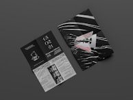 Two graphic print materials rest on a dark gray surface. One appears to be a folded booklet featuring bold red text and an abstract razor blade design with white and black wavy lines. The other is an unfolded brochure with information, elegant white typography against a black background, and illustrations, including event details and dates.