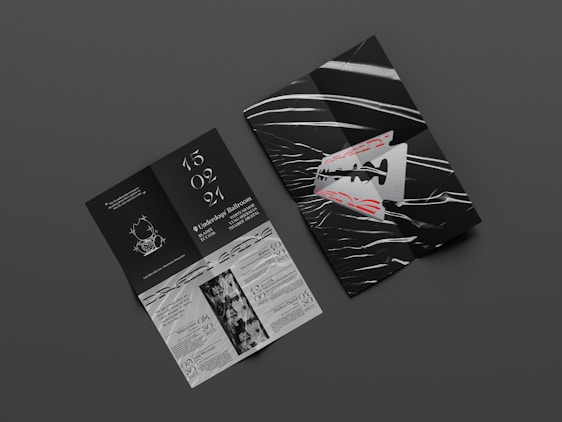 Two graphic print materials rest on a dark gray surface. One appears to be a folded booklet featuring bold red text and an abstract razor blade design with white and black wavy lines. The other is an unfolded brochure with information, elegant white typography against a black background, and illustrations, including event details and dates.