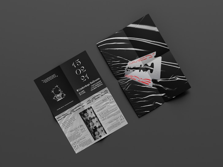 Two graphic print materials rest on a dark gray surface. One appears to be a folded booklet featuring bold red text and an abstract razor blade design with white and black wavy lines. The other is an unfolded brochure with information, elegant white typography against a black background, and illustrations, including event details and dates.