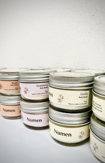 Several small glass jars with metal lids, each labeled as body butter with different fragrances such as vanilla and strawberry. The labels include instructions to store in a dark, cool place. The jars are neatly arranged and have a minimalistic design.