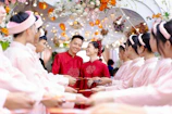 A couple dressed in red traditional attire stands smiling amid a ceremonial archway adorned with colorful flowers and lights. They are surrounded by a group of people in matching pink outfits, holding red envelopes in a celebratory setting.