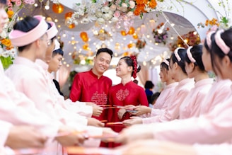 A couple dressed in red traditional attire stands smiling amid a ceremonial archway adorned with colorful flowers and lights. They are surrounded by a group of people in matching pink outfits, holding red envelopes in a celebratory setting.