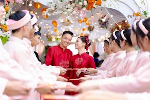A couple dressed in red traditional attire stands smiling amid a ceremonial archway adorned with colorful flowers and lights. They are surrounded by a group of people in matching pink outfits, holding red envelopes in a celebratory setting.