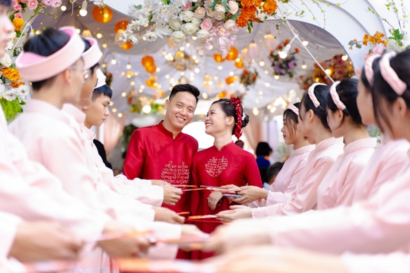 A couple dressed in red traditional attire stands smiling amid a ceremonial archway adorned with colorful flowers and lights. They are surrounded by a group of people in matching pink outfits, holding red envelopes in a celebratory setting.