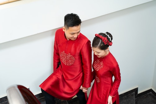 A couple dressed in traditional red attire is holding hands and walking up a staircase. The woman has her hair neatly styled with red floral accessories, and both have intricate embroidery on their garments.