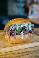 A delicious-looking sandwich with a sesame seed bun, filled with shredded meat, red onions, leafy greens, and drizzled with creamy and dark sauces. The sandwich is placed on a wooden surface, likely a cutting board.