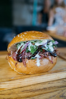 A delicious-looking sandwich with a sesame seed bun, filled with shredded meat, red onions, leafy greens, and drizzled with creamy and dark sauces. The sandwich is placed on a wooden surface, likely a cutting board.