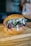 A delicious-looking sandwich with a sesame seed bun, filled with shredded meat, red onions, leafy greens, and drizzled with creamy and dark sauces. The sandwich is placed on a wooden surface, likely a cutting board.