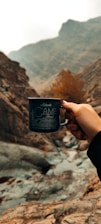 A hand holding a black camping mug against a background of rugged mountains. The terrain is rocky and desolate, with a muted, earthy palette. A patch of orange foliage provides a pop of color among the browns and grays.