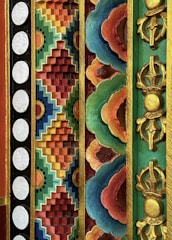 A vibrant and intricate design features geometric and floral patterns in a relief style with various colors and gold accents. The composition includes a series of repetitive designs with circular white dots on a black background, multicolored zigzag patterns, and ornate floral motifs with a gold outline. Deep reds, blues, greens, and oranges dominate the artwork, creating a rich and artistic impression.