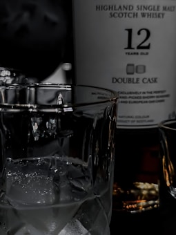 A glass filled with ice cubes sits in front of a bottle labeled as Highland Single Malt Scotch Whisky, aged 12 years, with 'Double Cask' mentioned. The image features contrasting lighting that highlights the clarity of the glass and the details on the bottle's label.
