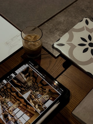 A tablet displaying an image of an interior setting with decorative elements lies on a wooden table. Next to it is a glass of iced coffee, a stack of papers, and a tile with a geometric pattern.