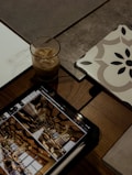 A tablet displaying an image of an interior setting with decorative elements lies on a wooden table. Next to it is a glass of iced coffee, a stack of papers, and a tile with a geometric pattern.