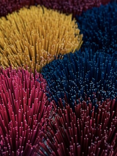 Clusters of tightly packed, vertical, colorful plastic bristles are arranged closely together. The colors of the bristles include yellow, blue, and shades of pink or red. The bristles create a visually stimulating, almost abstract pattern due to their density and varying lengths.