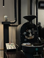 A coffee roasting machine with a control panel is situated in an industrial setting. The machine is black with various knobs and metallic components, and a cooling tray is visible in the foreground.