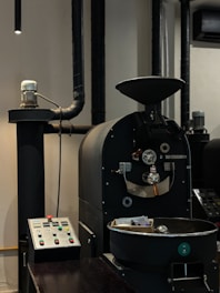 A coffee roasting machine with a control panel is situated in an industrial setting. The machine is black with various knobs and metallic components, and a cooling tray is visible in the foreground.
