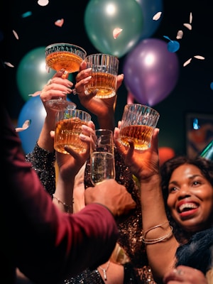 A group of people raise their glasses in a celebratory toast, with colorful balloons in the background. Confetti is scattered through the air, adding to the festive atmosphere.