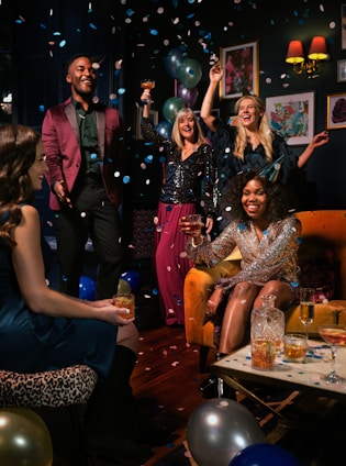 A lively celebration with a group of people joyfully raising their glasses. Multicolored confetti and balloons enhance the festive atmosphere in a dimly lit room adorned with framed art.