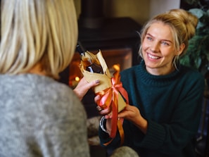 A smiling woman with blond hair is handing a wrapped gift, possibly a bottle, to another person. The gift is decorated with a red ribbon. They are indoors, in front of a fireplace that is producing a warm, cozy atmosphere.
