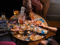 A table filled with a variety of drinks, including whiskey and cocktails, surrounded by people. The table is decorated with colorful confetti and a glass decanter filled with an amber liquid. The scene has a festive atmosphere with a focus on celebration.