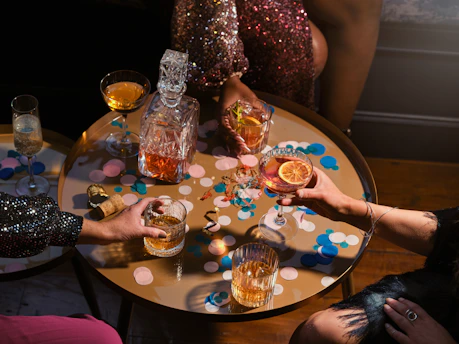 A table filled with a variety of drinks, including whiskey and cocktails, surrounded by people. The table is decorated with colorful confetti and a glass decanter filled with an amber liquid. The scene has a festive atmosphere with a focus on celebration.
