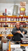 A man is packaging goods in a shop filled with shelves of various spices and herbs stored in jars and bags. The shelves are neatly organized, displaying an array of colorful containers. The man appears to be focused on his task, standing behind a counter with a digital scale.
