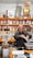 A man is packaging goods in a shop filled with shelves of various spices and herbs stored in jars and bags. The shelves are neatly organized, displaying an array of colorful containers. The man appears to be focused on his task, standing behind a counter with a digital scale.