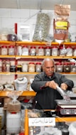 A man is packaging goods in a shop filled with shelves of various spices and herbs stored in jars and bags. The shelves are neatly organized, displaying an array of colorful containers. The man appears to be focused on his task, standing behind a counter with a digital scale.