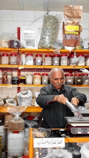A man is packaging goods in a shop filled with shelves of various spices and herbs stored in jars and bags. The shelves are neatly organized, displaying an array of colorful containers. The man appears to be focused on his task, standing behind a counter with a digital scale.