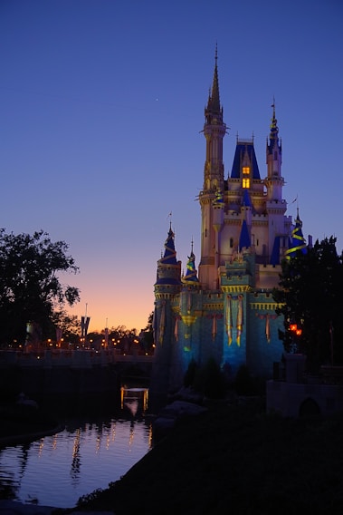 A fairytale-like castle is illuminated against a twilight sky. The architecture features tall spires and intricate details, with some areas glowing in soft lighting. A calm body of water reflects the structure, surrounded by trees and landscape.